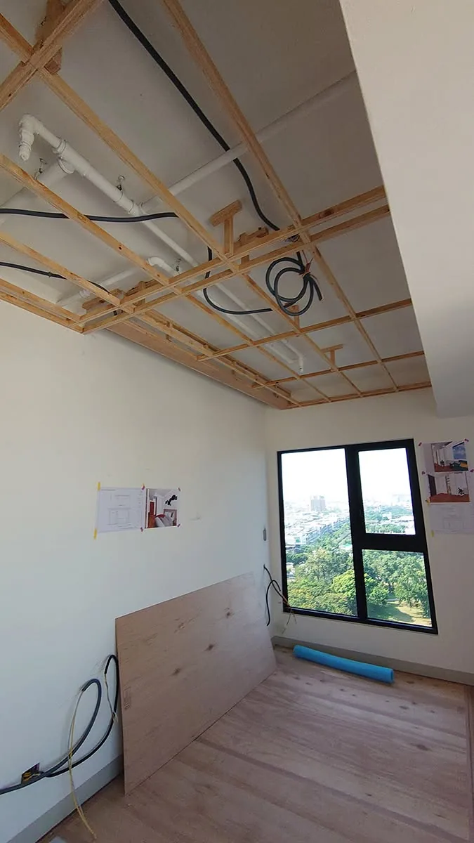 臥室天花板角材骨架間可見白色排水管與黑色電線，牆面張貼設計圖